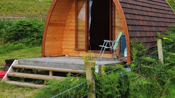 A small, wooden, arched cabin with a wooden deck and a ladder, surrounded by greenery and hills. - Canna Campsite (Isle of Canna)