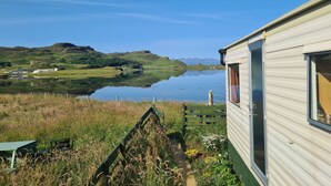 A lakeside view with a mobile home, a picnic table, and a grassy area. - Canna Campsite (Isle of Canna)