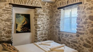 A stone-walled room with a bed, a painting, and a window.