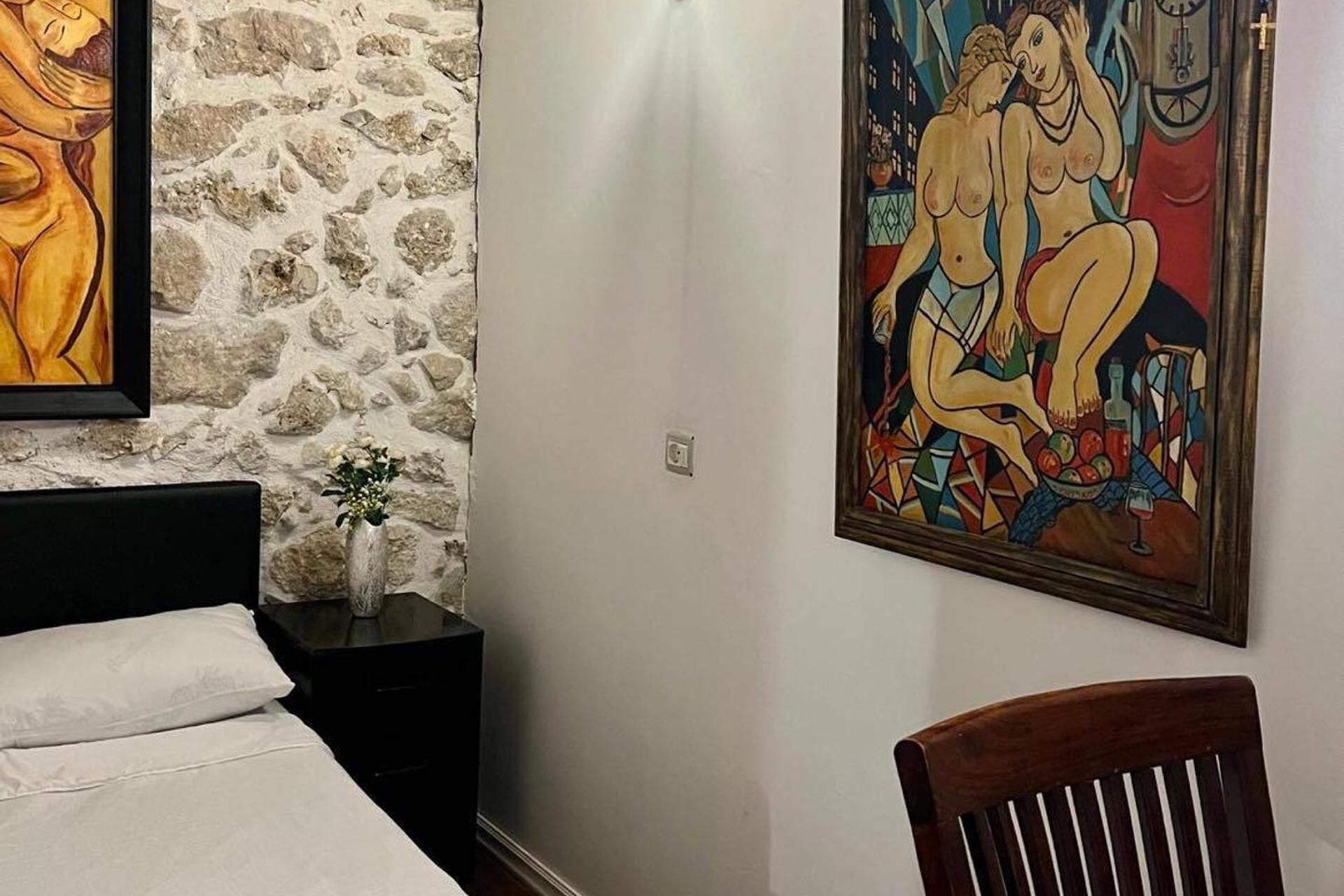 A bedroom with a stone wall, a bed with white linens, a nightstand with a vase of flowers, and a painting on the wall.