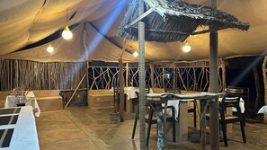 A tented dining area with tables and chairs, a thatched roof, and hanging light bulbs.