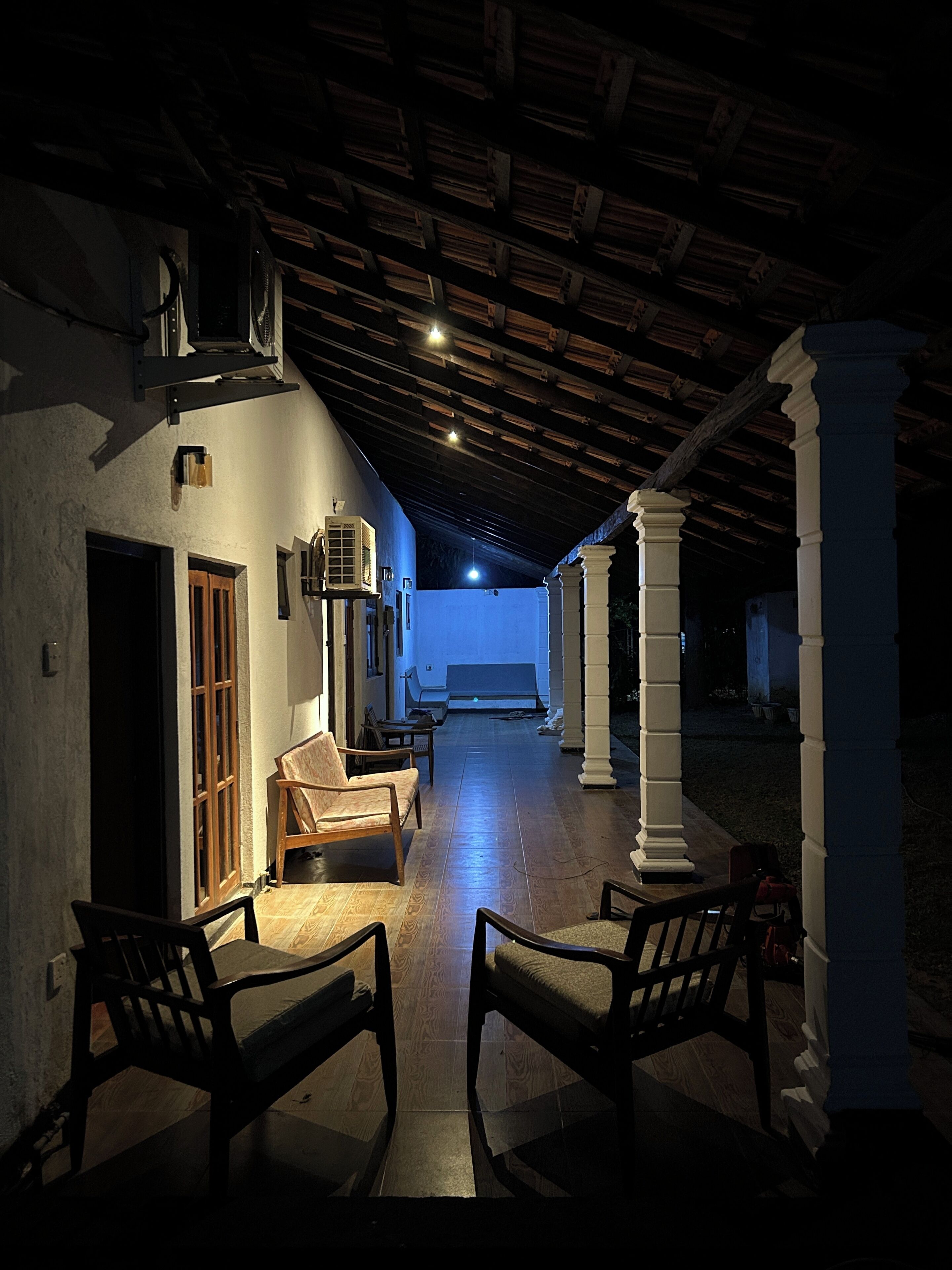 Un pasillo tenuemente iluminado con suelo de madera, un banco y vista a una habitación con una ventana iluminada en azul.