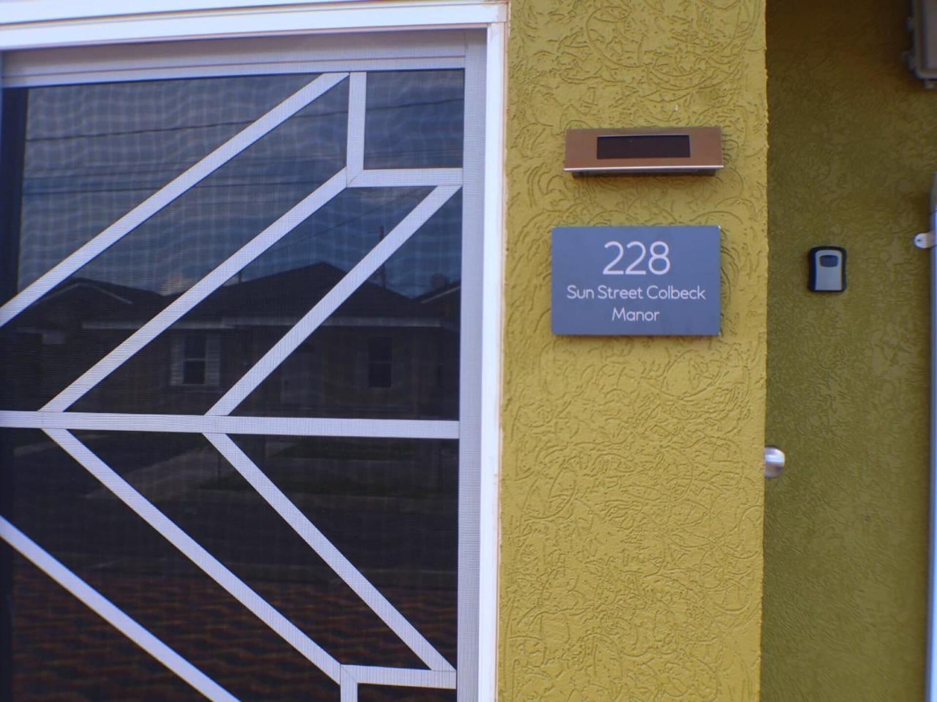 A building entrance with a large glass door featuring a geometric pattern. A sign above the door displays the number 228 and the name 'Sun Street Colbeck Manor'.