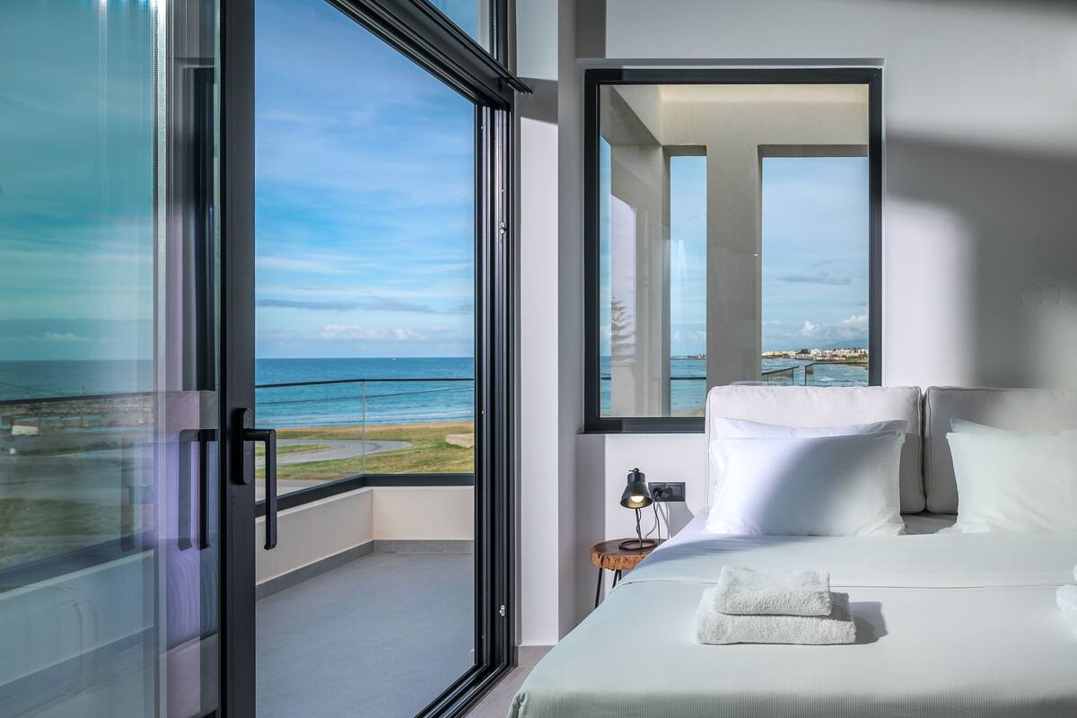 A modern bedroom with a large window overlooking a beach, a bed with white linens, and a small bedside table with a lamp.