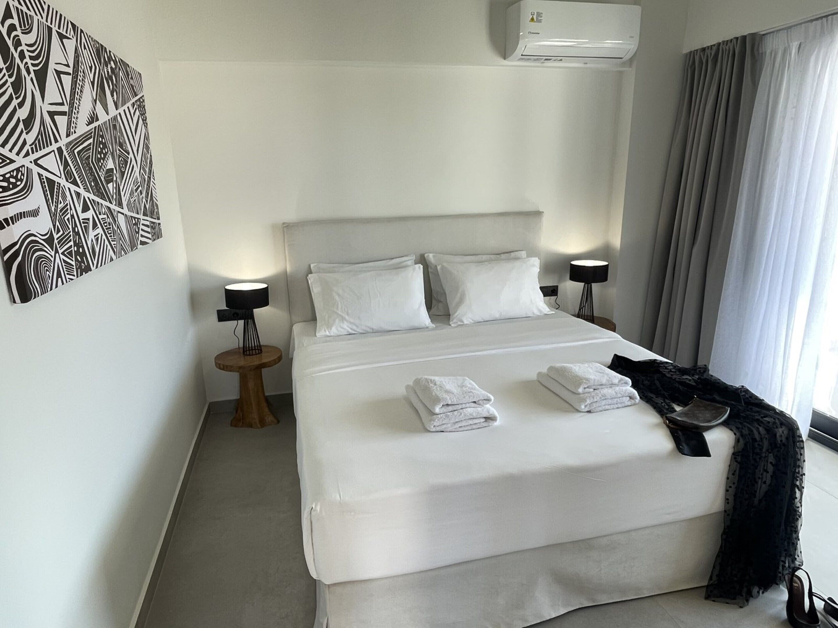 A hotel room with a large bed, two bedside tables with lamps, a black and white abstract artwork on the wall, and a window with curtains.