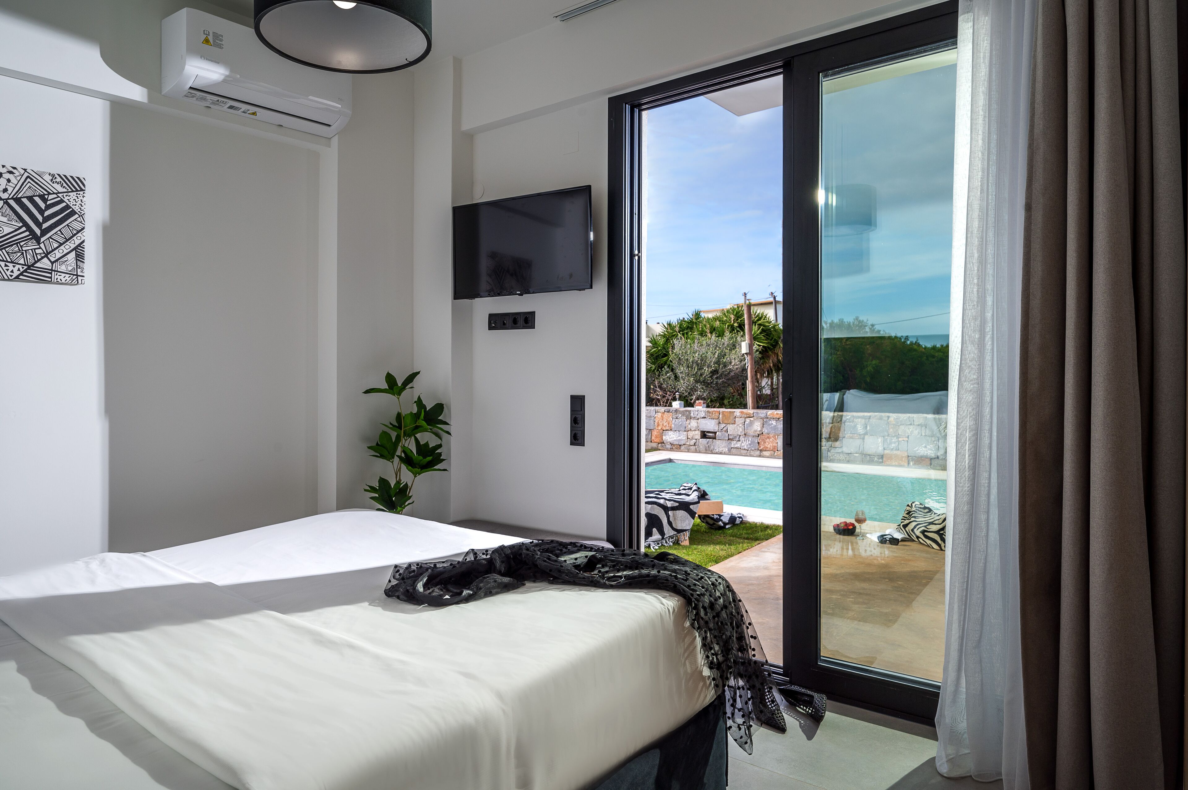 Une chambre avec un lit, un téléviseur fixé au mur et une porte-fenêtre coulissante donnant sur une zone avec piscine.