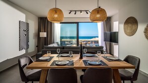 A dining area with a wooden table set for six, two hanging wicker lamps, a large window with a view of the sea, and a flat-screen TV mounted on the wall. - Destino Luxury Villas Gournes (Hersonissos)
