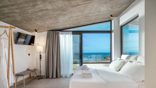 A modern hotel room with a large bed, a bench, a TV, and a view of the ocean. - Destino Luxury Villas Gournes (Hersonissos)