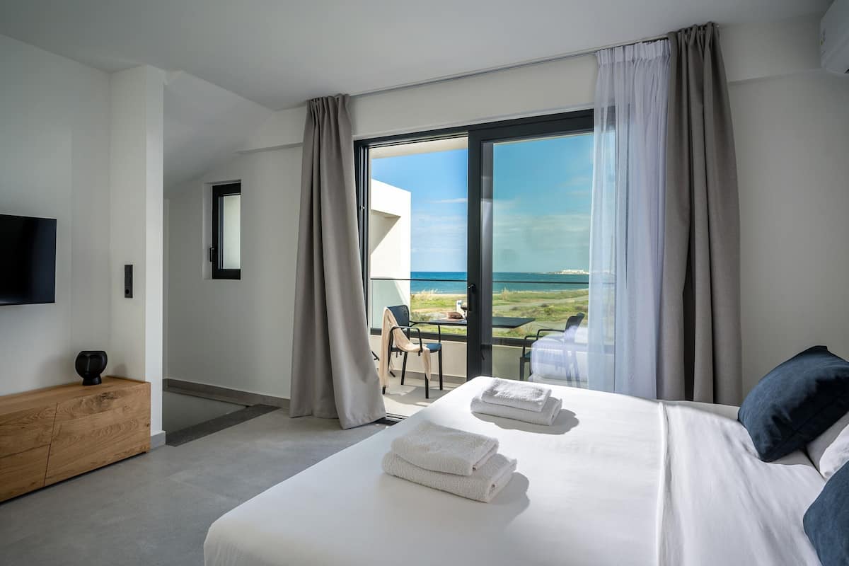 A modern hotel room with a large bed, a wooden side table, a flat-screen TV, and a balcony with a view of the sea.