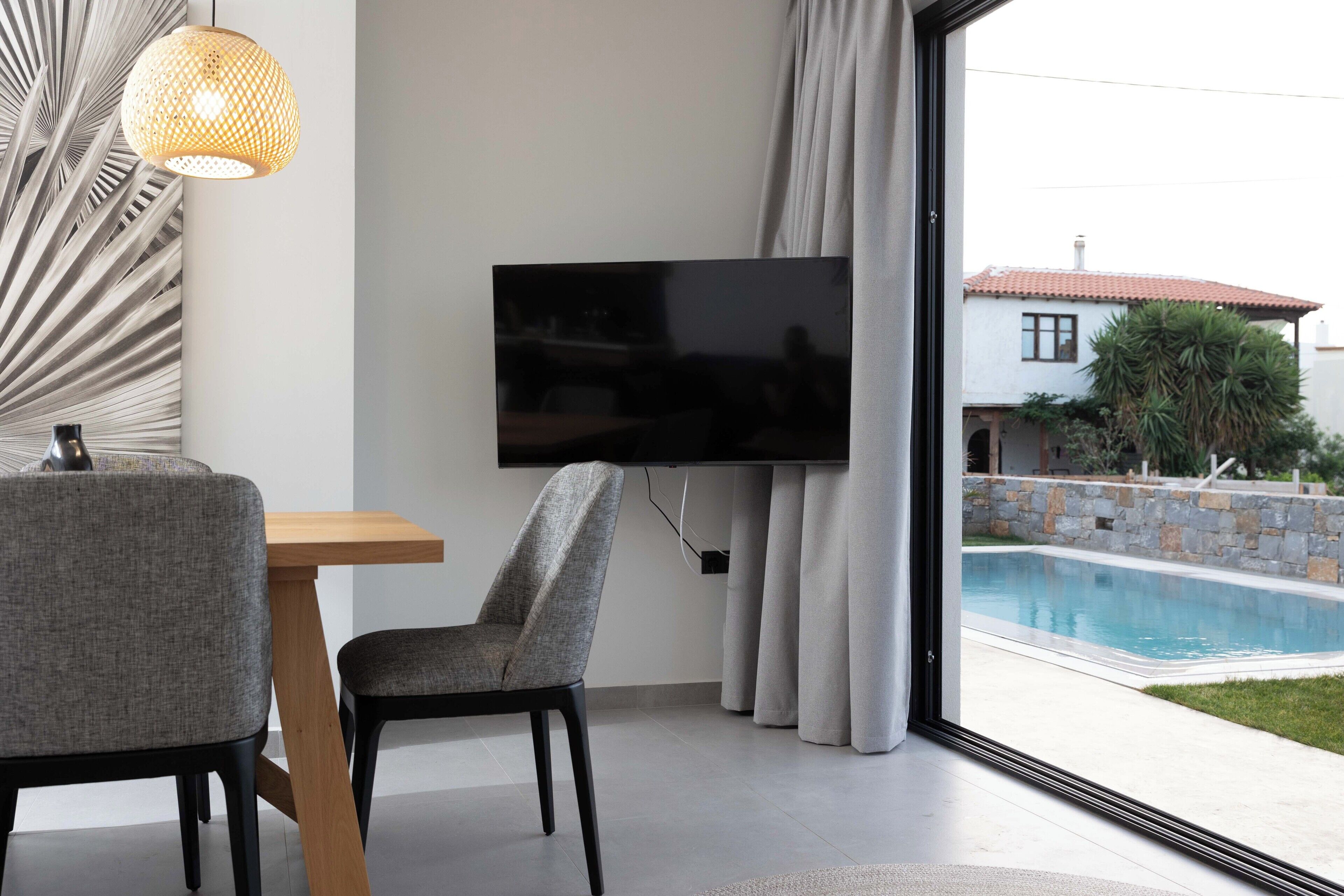 A modern living room with a flat-screen TV mounted on the wall, a dining table with two chairs, and a view of a swimming pool outside.
