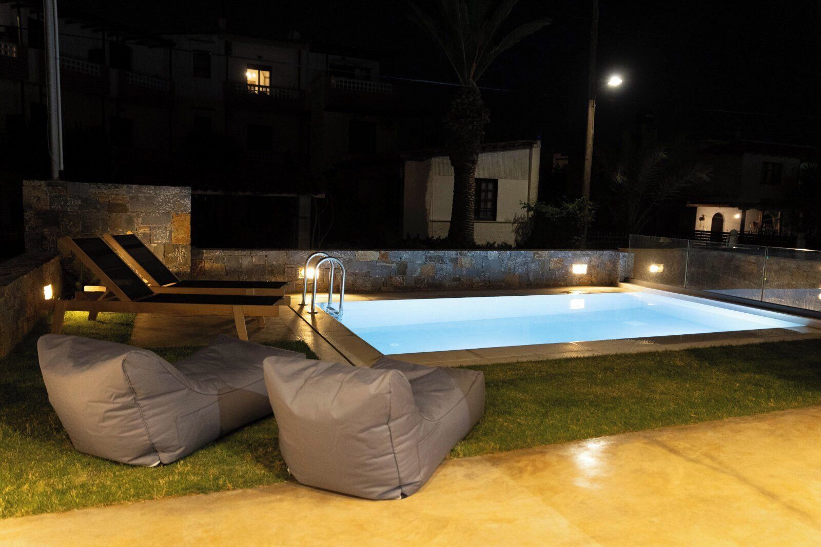 A nighttime swimming pool area with a lounge chair and two bean bags, surrounded by stone walls and a well-lit pool.
