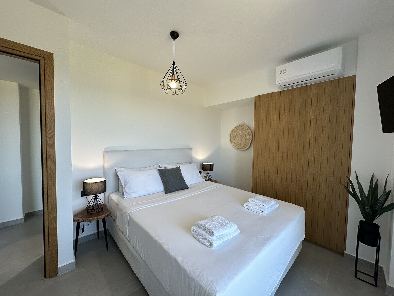 A modern hotel room with a large bed, a bedside table with a lamp, an air conditioning unit, and a potted plant.