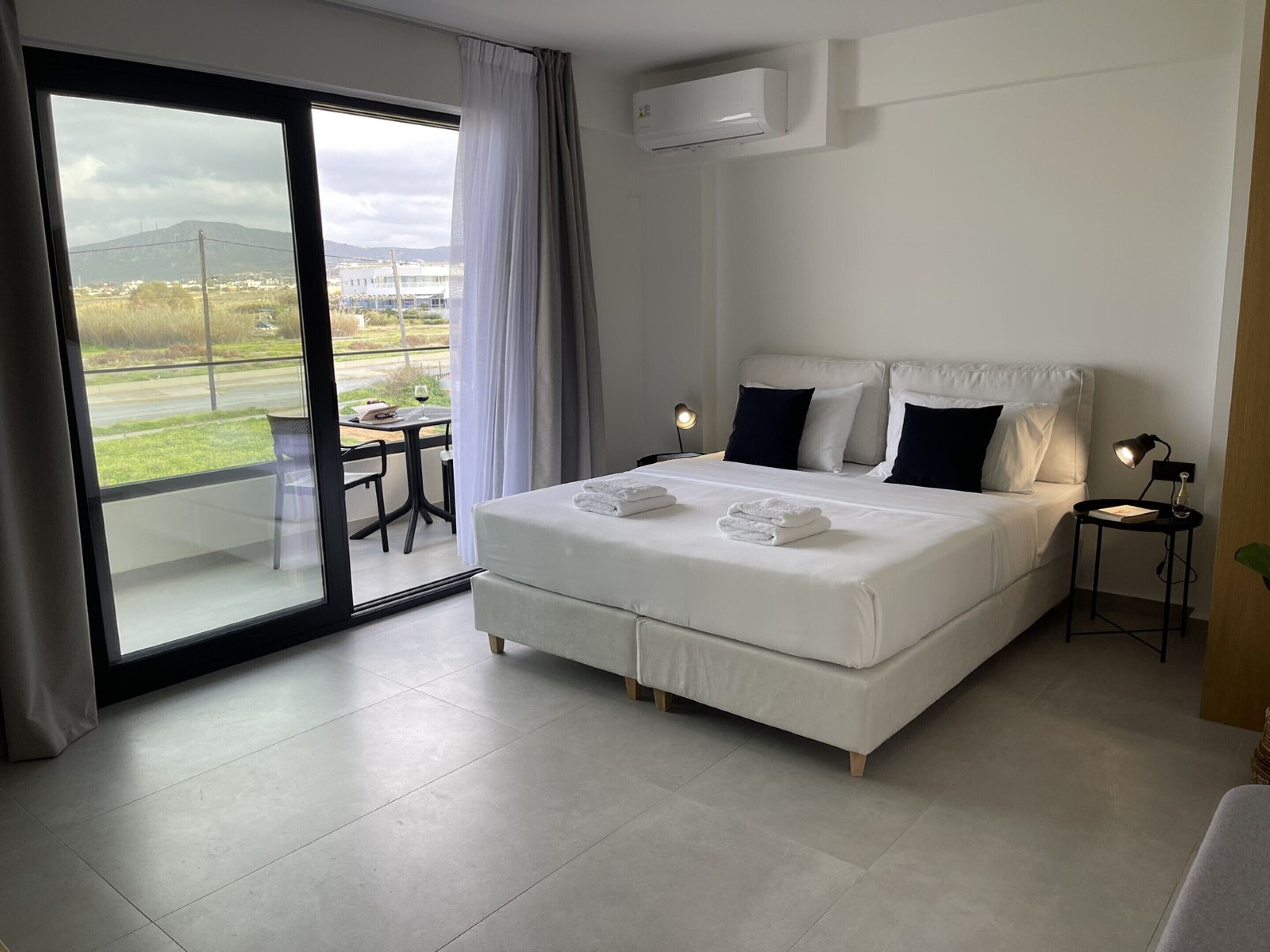 A modern hotel room with a large bed, a balcony with a table and chairs, and an air conditioning unit on the wall.