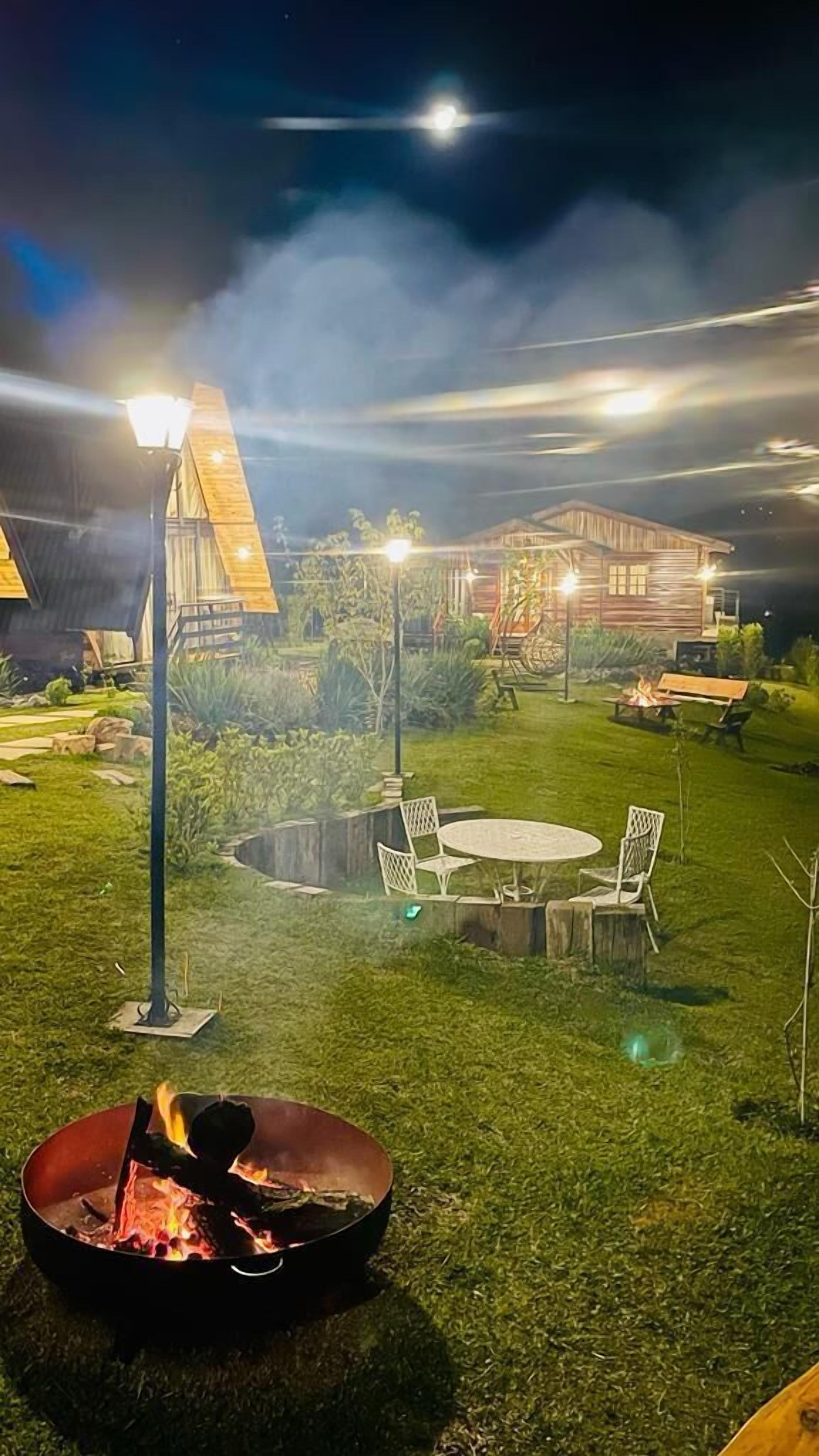 Área externa noturna com lareira, assentos e um edifício ao fundo.