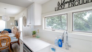 A modern kitchen with a white countertop, a sink, and a window with a view of trees. There is a sign above the window that reads "Welcome to Lucky Enough." - 4 BDR, Ocean Front Condo (Lucky Enough) in Ocean Grove Complex (Salter Path)