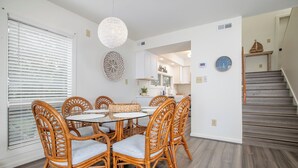 A dining area with wicker chairs and a glass table, a kitchen with white cabinets, and a wooden staircase. - 4 BDR, Ocean Front Condo (Lucky Enough) in Ocean Grove Complex (Salter Path)