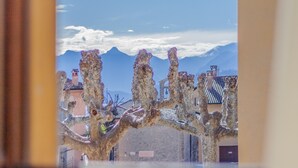 A view of a tree with unique, twisted branches in front of a building with a tiled roof and mountains in the background. - Miranda's Home (Varenna)
