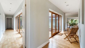 Um corredor amplo com piso de madeira, grandes portas de vidro deslizantes que dão para uma área externa e um conjunto de jantar com cadeiras de madeira.