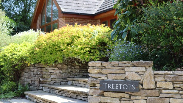 A stone wall with a sign reading "Treetops" and a house with a sloped roof and large windows.