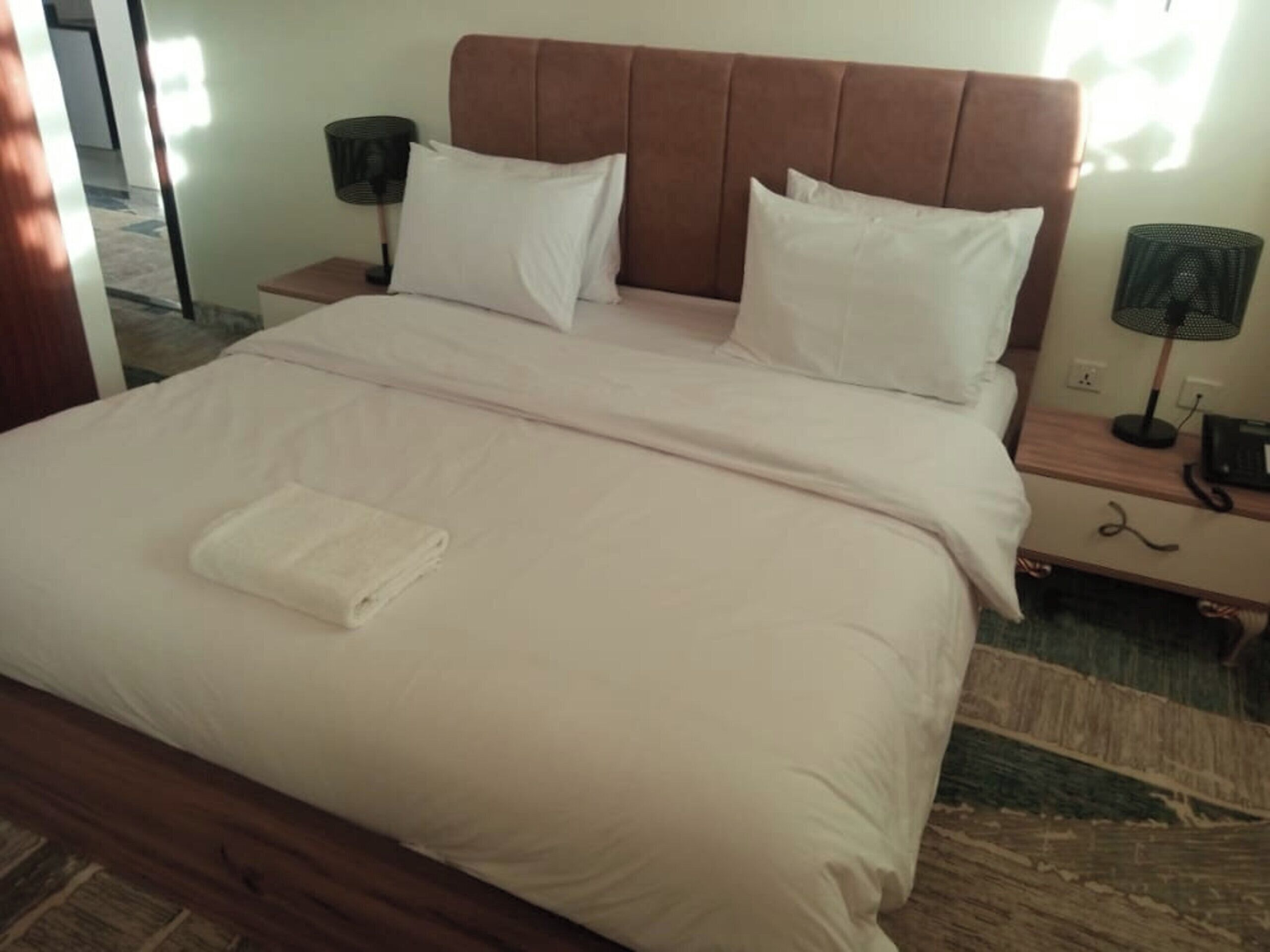A neatly made bed with a brown headboard, white pillows, and a folded white towel on the footboard. There are two bedside tables with lamps and a phone.