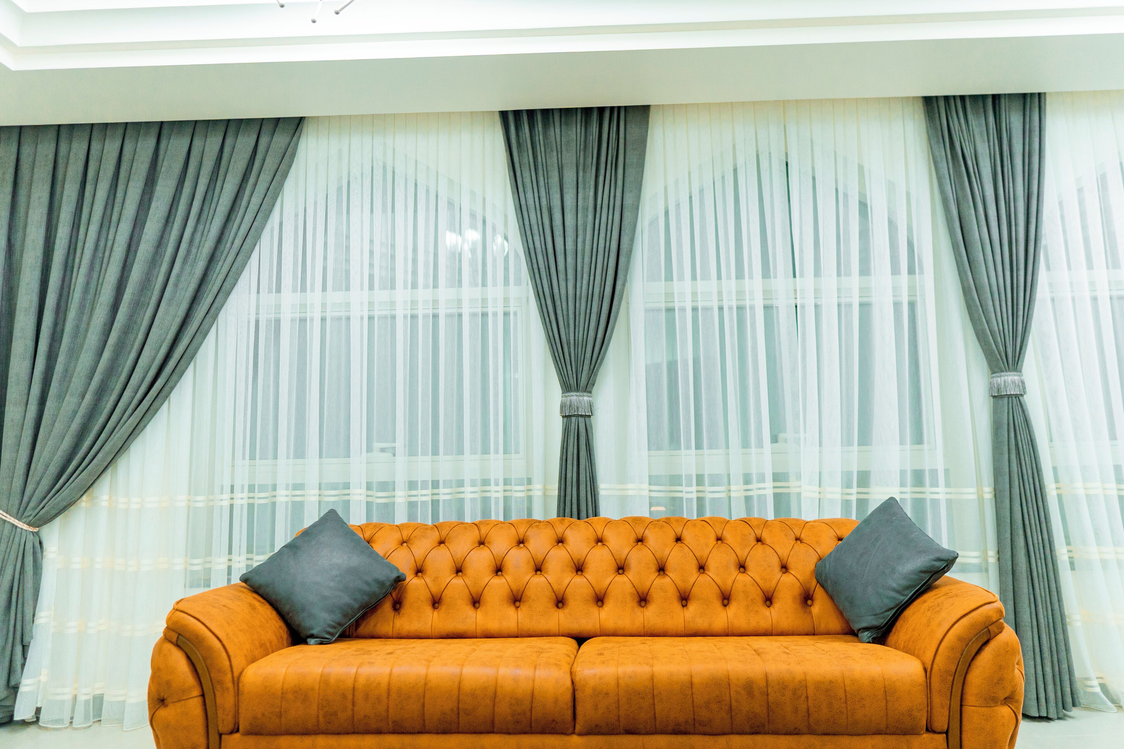 A room with a tufted orange sofa, grey curtains, and sheer white drapes.