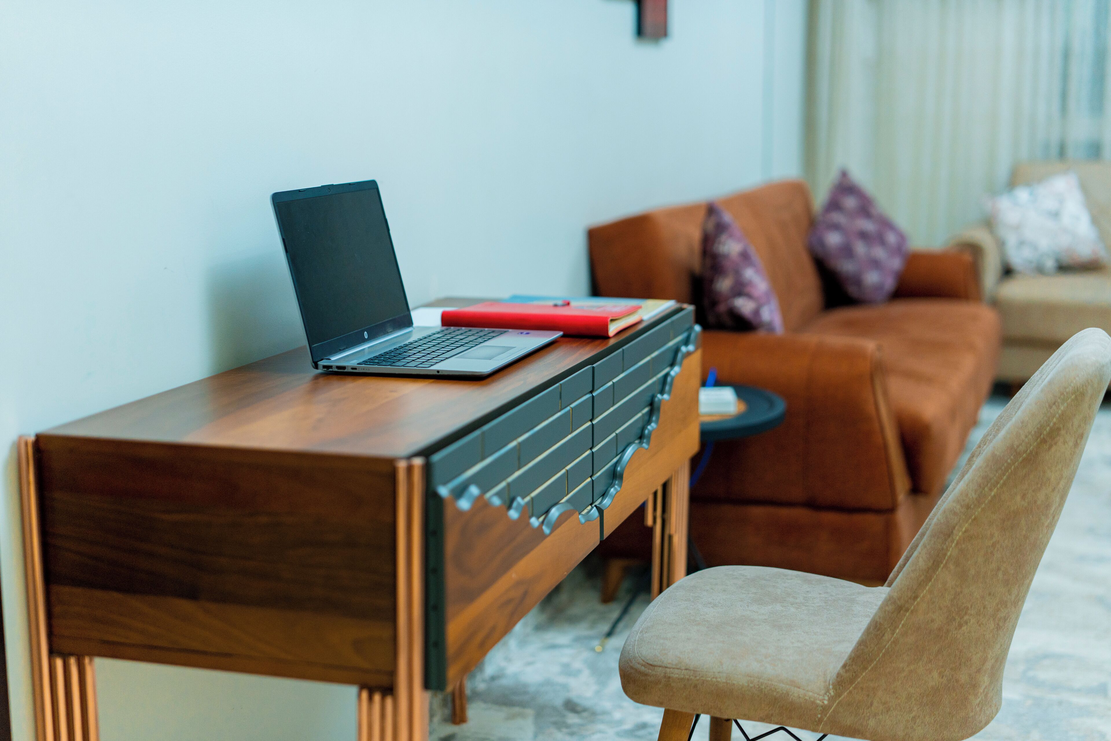 Un bureau en bois avec un ordinateur portable et un livre rouge, un canapé marron avec des coussins à motifs et un fauteuil beige dans un salon.