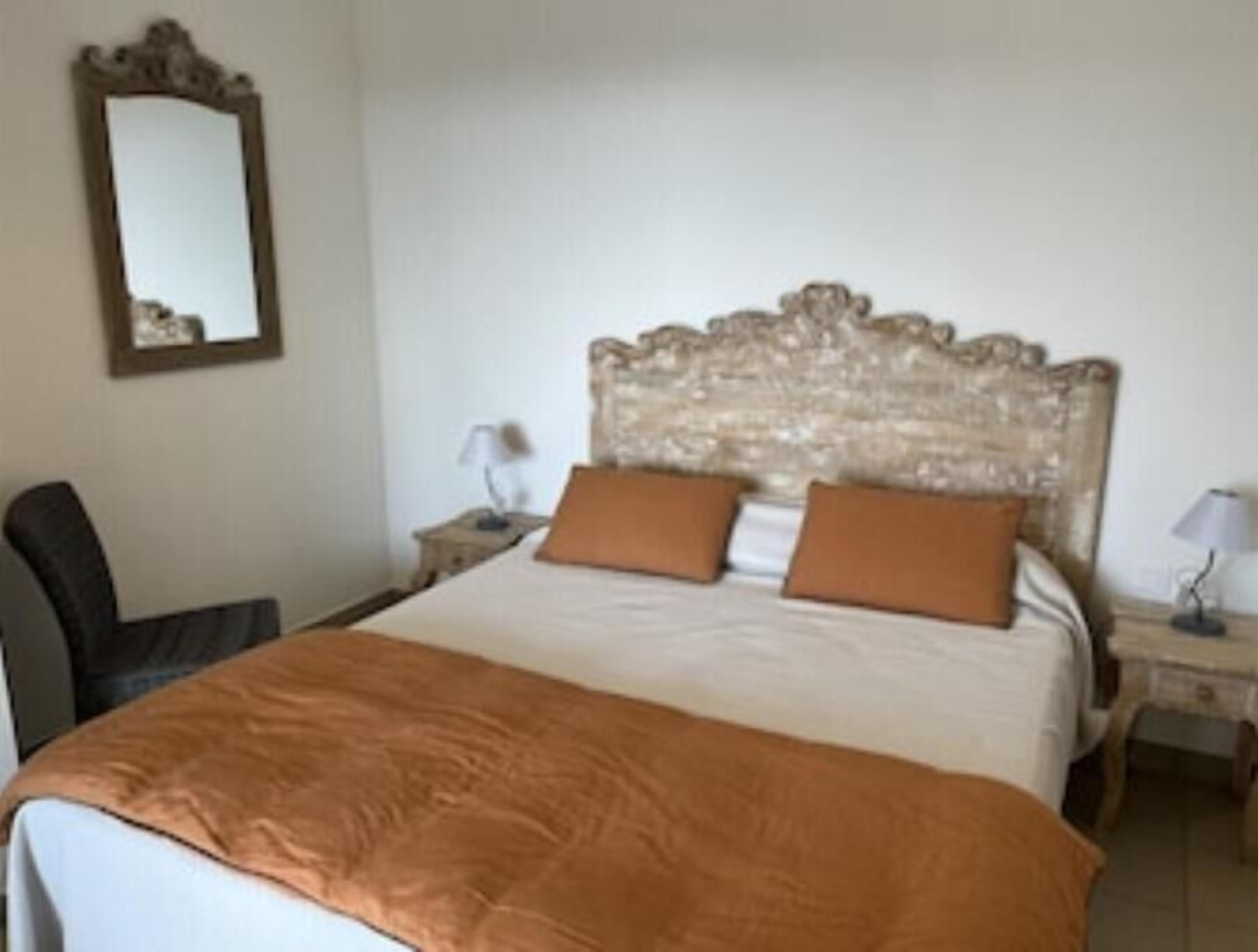 A hotel room with a large bed, two brown pillows, a wooden headboard, a nightstand with a lamp, and a chair.