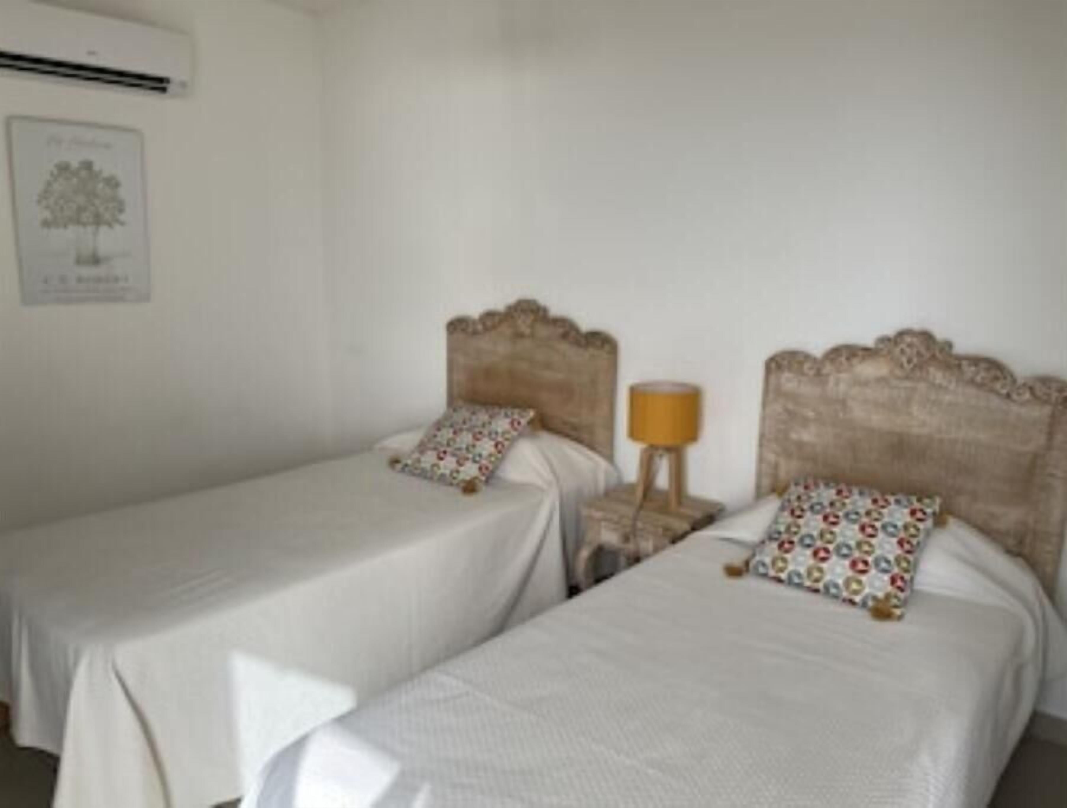 Ein Zimmer mit zwei Einzelbetten, jeweils mit einem Holz-Kopfende und passendem Kissen. Dazwischen steht ein kleiner Nachttisch mit einer Lampe.