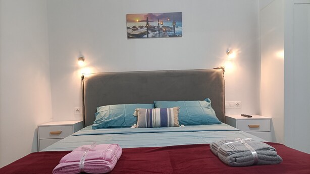 A neatly made bed with a red blanket and two sets of towels, a nightstand with a remote control, and a wall-mounted picture of a sunset over the sea. - City Aurora (Athens)