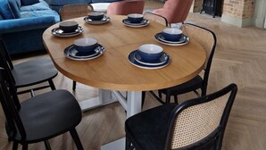 Un coin repas avec une table ronde en bois, dressée pour six personnes, entourée de chaises noires à assise tissée. Un canapé bleu et un fauteuil rose se trouvent à l’arrière-plan.