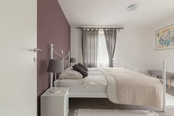 Une chambre moderne avec un lit blanc, une table de chevet avec une lampe, une fenêtre avec des rideaux et une œuvre d’art accrochée au mur.