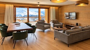 A modern living room with a wooden dining table, green chairs, a sofa, and a television mounted on the wall.