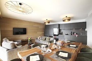 Uma sala de estar moderna com parede de madeira, sofás cinzas, uma mesa de jantar posta para quatro pessoas e uma cozinha com armários escuros e uma televisão de tela plana.