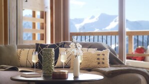 Une pièce chaleureuse avec un décor intérieur en bois, un canapé agrémenté de coussins à motifs, une petite table avec une bougie, un vase rempli de fleurs et une vue sur des montagnes enneigées.