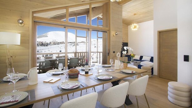 Un espace repas avec une longue table dressée pour un repas, de grandes fenêtres offrant une vue sur un paysage enneigé et un coin salon confortable avec un canapé et des plantes décoratives.