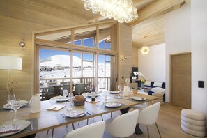 A dining area with a long table set for a meal, large windows offering a view of a snowy landscape, and a cozy seating area with a sofa and decorative plants. - 10m from ski slopes, 3rd floor, fitness, swimming pool, sauna, hammam, balcony, tv, ski locker, 93m² (Huez)