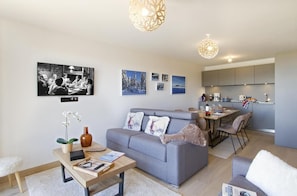 A modern living room with a grey sofa, a wooden coffee table, and a dining area with a kitchen in the background. - Ground floor, fitness, swimming pool, sauna, hammam, terrace, tv, ski locker, 55m², Alpe d'Huez (Huez)