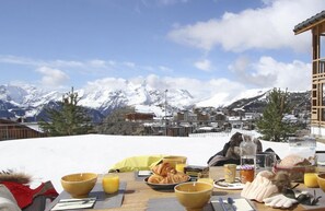 Mesa de café da manhã posta com croissants, suco de laranja e uma garrafa de água, com vista para uma paisagem montanhosa coberta de neve.