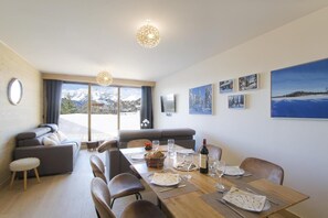 A modern dining area with a wooden table set for six, a sofa, and a view of mountains through large windows. - Ground floor, fitness, swimming pool, sauna, hammam, terrace, tv, ski locker, 55m², Alpe d'Huez (Huez)