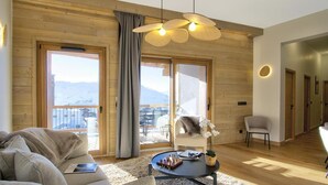Amplia sala de estar con piso de madera, un sofá, una mesa de centro con piezas de ajedrez y vistas a un balcón con el paisaje de montañas al fondo.
