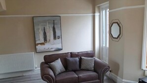 Un salon avec un canapé en cuir marron, un tableau représentant des bouteilles accroché au mur et une fenêtre ornée d’un miroir aux motifs géométriques.