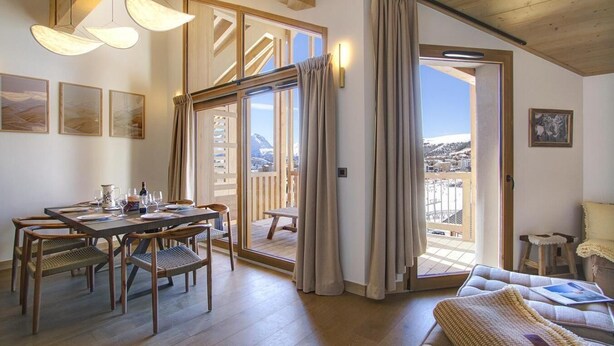 Une pièce chaleureuse avec une table à manger en bois, des chaises et un canapé. Une porte-fenêtre coulissante donne accès à un balcon extérieur d’où l’on peut admirer la vue sur les montagnes.