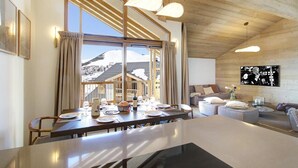 Un coin repas avec une grande table dressée pour un repas, un canapé et un téléviseur, dans une pièce au plafond en bois offrant une vue sur un paysage enneigé.