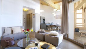 Un salon moderne avec un canapé, un pouf et une table basse, un coin repas avec des meubles en bois, et une cuisine en arrière-plan.