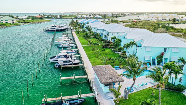 Un port de plaisance avec des bateaux amarrés, un espace piscine bordé de palmiers et une rangée de bâtiments d’un bleu clair.
