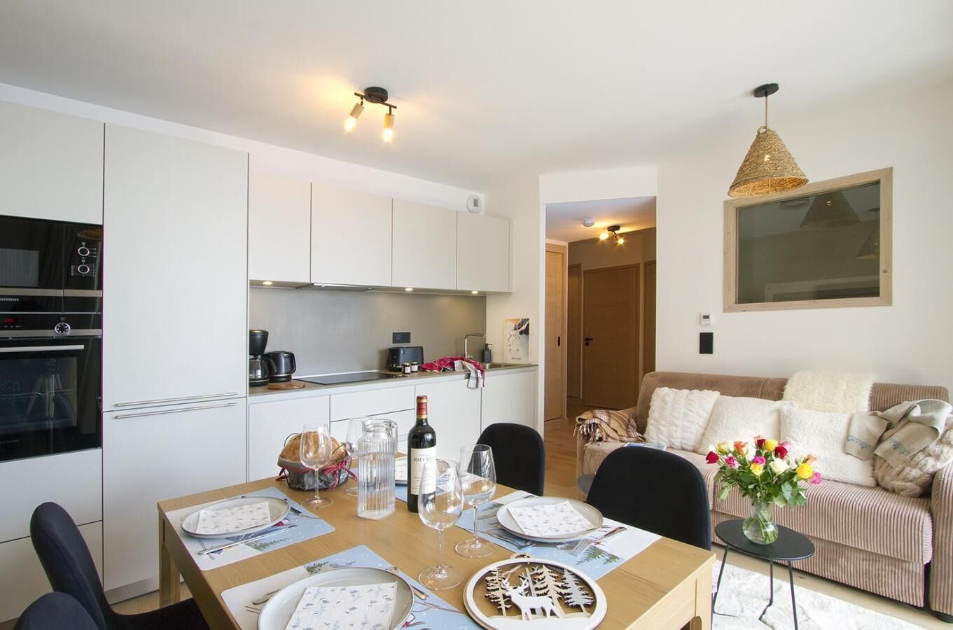 Une cuisine moderne avec des meubles blancs, une table à manger dressée pour quatre personnes et un coin salon accueillant avec un canapé et un vase de fleurs.