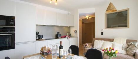 Une cuisine moderne avec des meubles blancs, une table à manger dressée pour quatre personnes et un coin salon accueillant avec un canapé et un vase de fleurs.