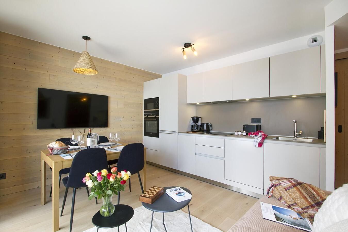 A modern kitchen with white cabinets, a flat-screen TV, and a dining area with a wooden table and chairs.