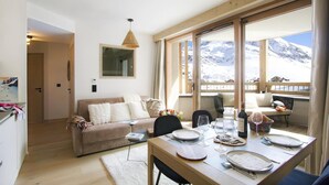 Un coin repas avec une table dressée pour deux personnes, un canapé et une grande fenêtre donnant sur les montagnes.