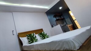 Un dormitorio con una cama, cabecera de madera, pared blanca y una pequeña zona de kitchenette con un lavamanos y armarios.